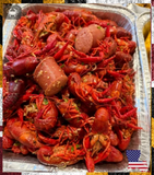A large aluminum tray filled with cooked crawfish and sausage, photographed from above. The crawfish are bright red-orange, mixed with sliced sausage pieces and visible green onions and garlic pieces.