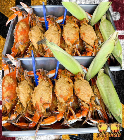 Two metal trays filled with cooked blue crabs topped with a golden-brown seasoning mix, garnished with green corn husks, and held together by blue plastic picks. A 'sally beach' logo is visible in the bottom right corner.