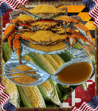 A plated seafood spread featuring several cooked crabs topped with yellow seasoning, fresh corn on the cob, a silver serving dish, and a small bowl of brown dipping sauce, set against a decorative American flag and checkered border.