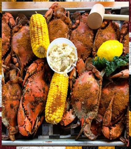 A top-down view of a seafood boil platter featuring cooked crabs seasoned with spices, two ears of corn, a small cup of creamy butter, a lemon wedge, and green garnish on a tray.