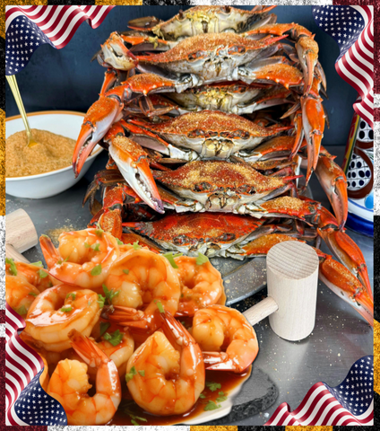 A large stack of cooked blue crabs on a dark serving platter, with a plate of cooked shrimp in sauce in the foreground and small American flags around the edges.